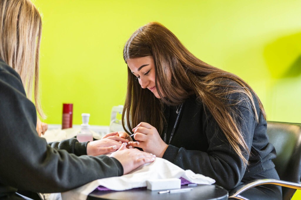 A young woman with long brown hair receiving a manicure from a student in a nail care class at Suffolk New College, with a bright green wall in the background.