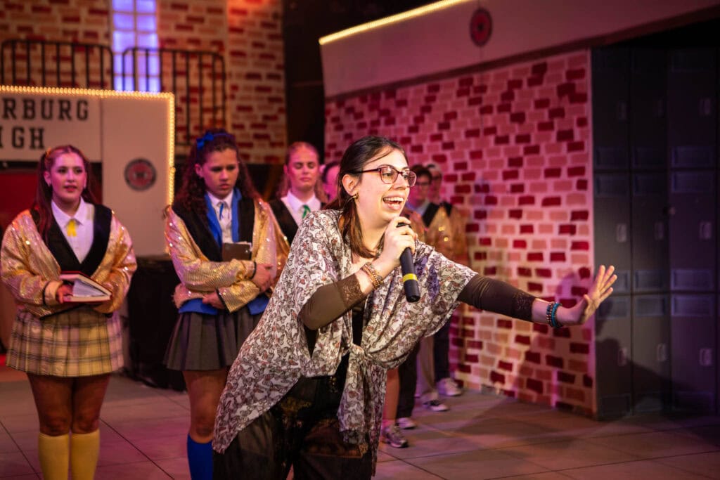 Group of students at Suffolk New College during a performance, with one girl singing into a microphone and others in school uniforms, showcasing college activities and student engagement.