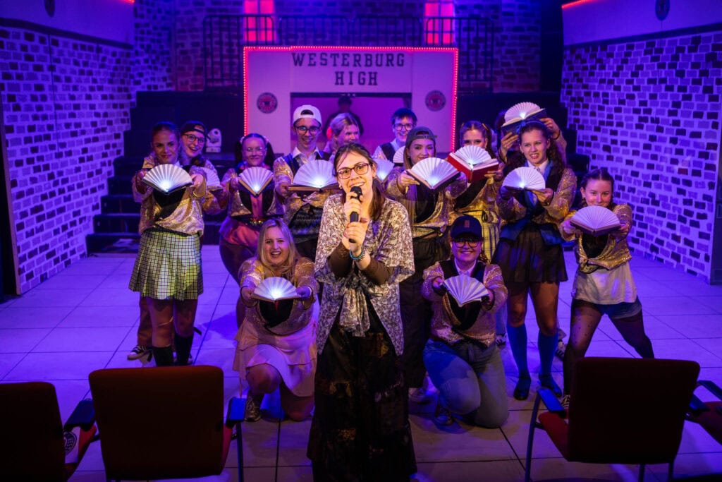 Group of students performing on stage at Suffolk New College, with a teacher or director in front, under vibrant purple lighting, showcasing arts and drama education.
