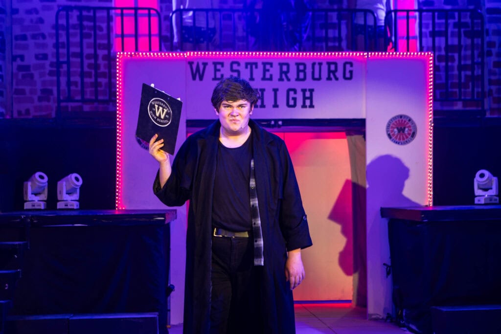 A student at Suffolk New College standing on stage during a performance, holding a script or booklet, with a backdrop displaying "Westerburg High" and stage lighting, showcasing college arts programs.