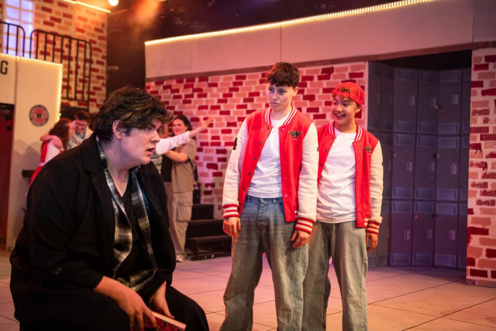 Group of Suffolk New College students performing a play on stage, with one student sitting and two others standing, in a theatre setting with brick walls and stage lighting.