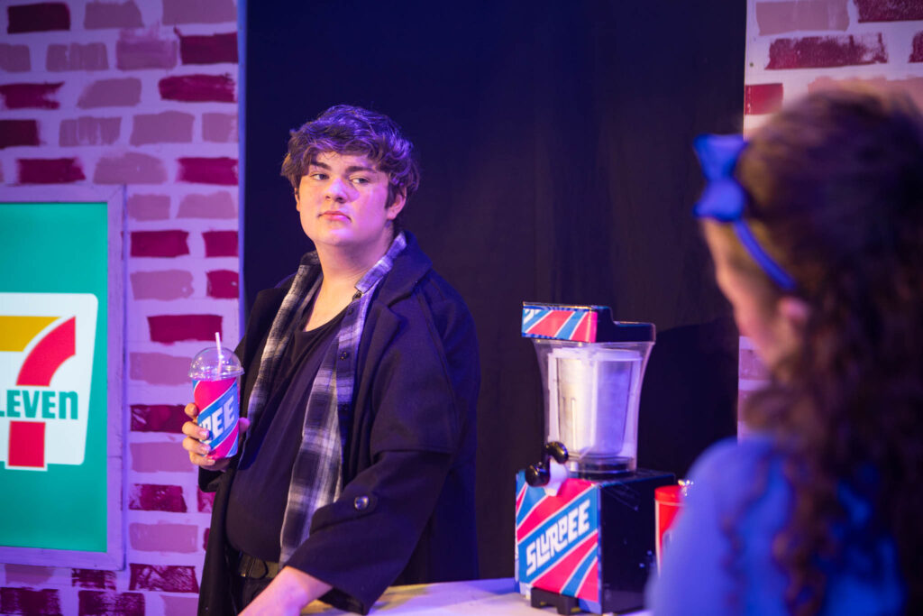 Young male student at Suffolk New College participating in a 7-Eleven promotional event, holding a drink, with a brick wall background and a 7-Eleven branded setup.