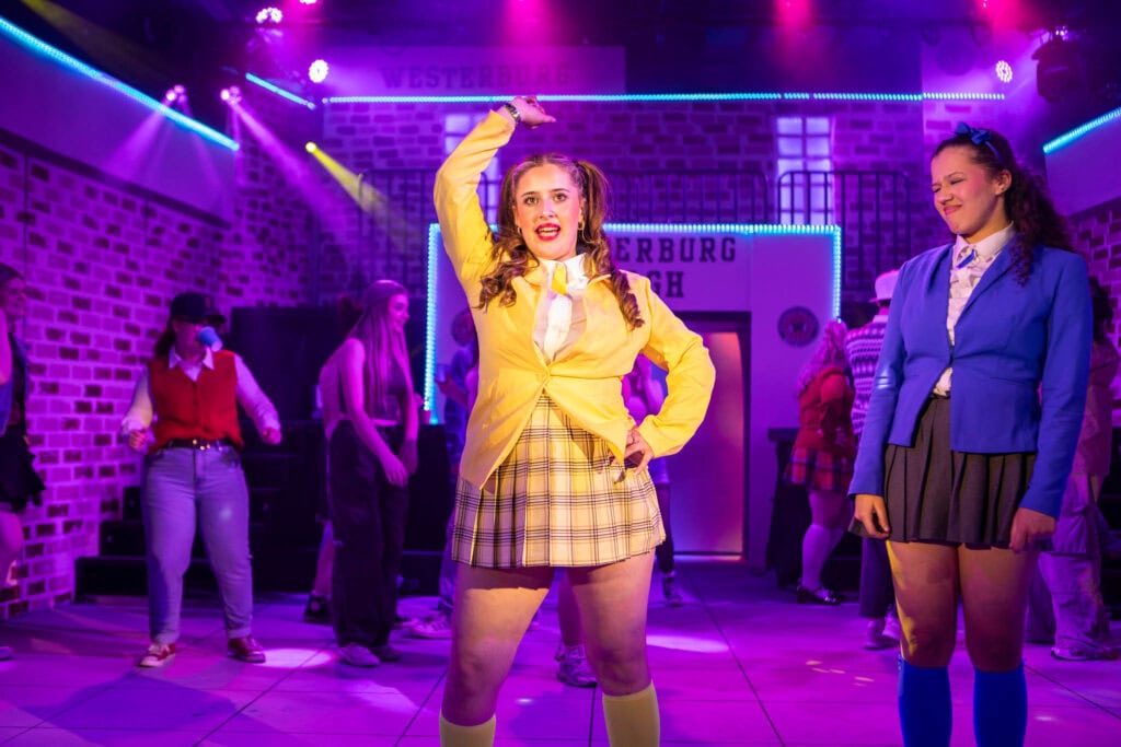 A vibrant scene from a theatre production at Suffolk New College, featuring students in colourful costumes performing on stage with dynamic lighting and a brick wall backdrop.
