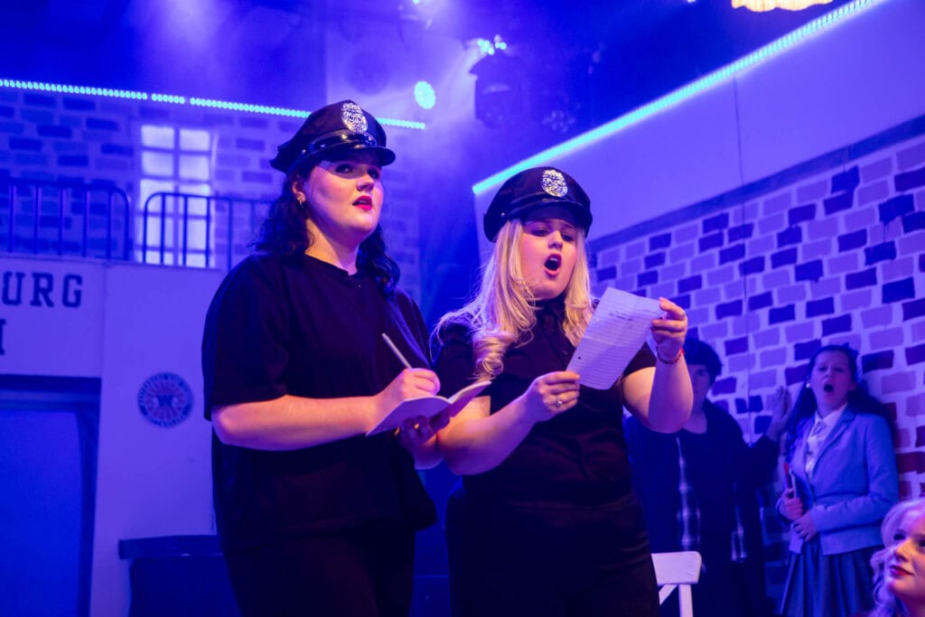 Two women dressed as police officers participating in a training or role-play activity at Suffolk New College, with other students and staff in the background, under vibrant blue lighting.