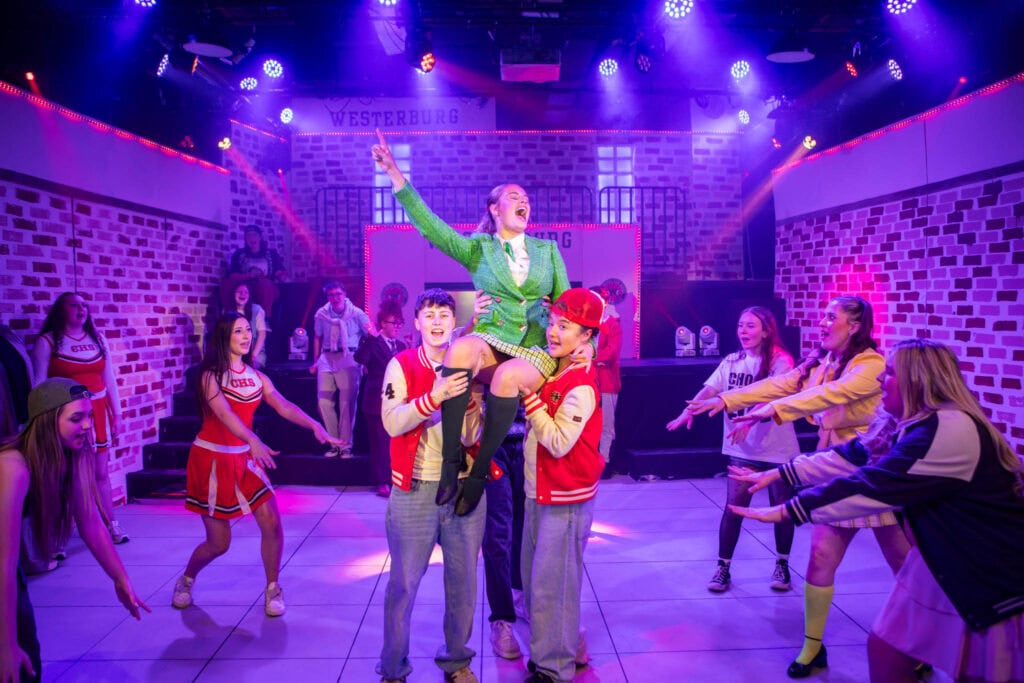 An energetic theatre scene at Suffolk New College featuring students in colourful costumes performing on stage with vibrant lighting.
