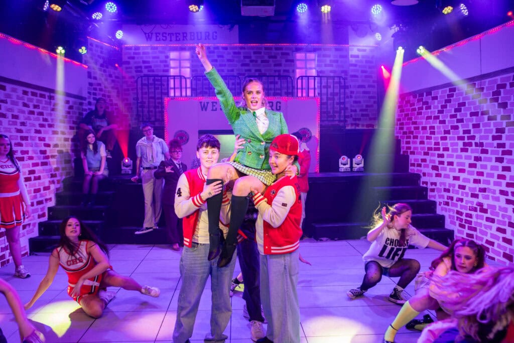 Group of young performers on stage at Suffolk New College, with a woman lifting a child, surrounded by other children in colorful costumes, under vibrant stage lighting.
