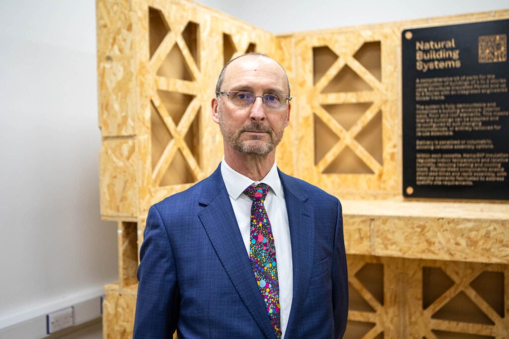 Man in formal suit standing in front of a wooden display about natural building systems at Suffolk New College, promoting construction and sustainable building skills.