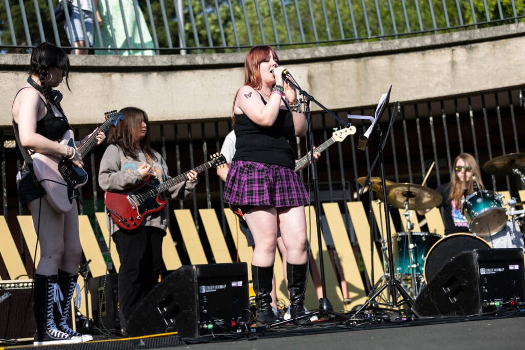 Group of young female students performing live music on an outdoor stage at Suffolk New College, showcasing student talent and college events.