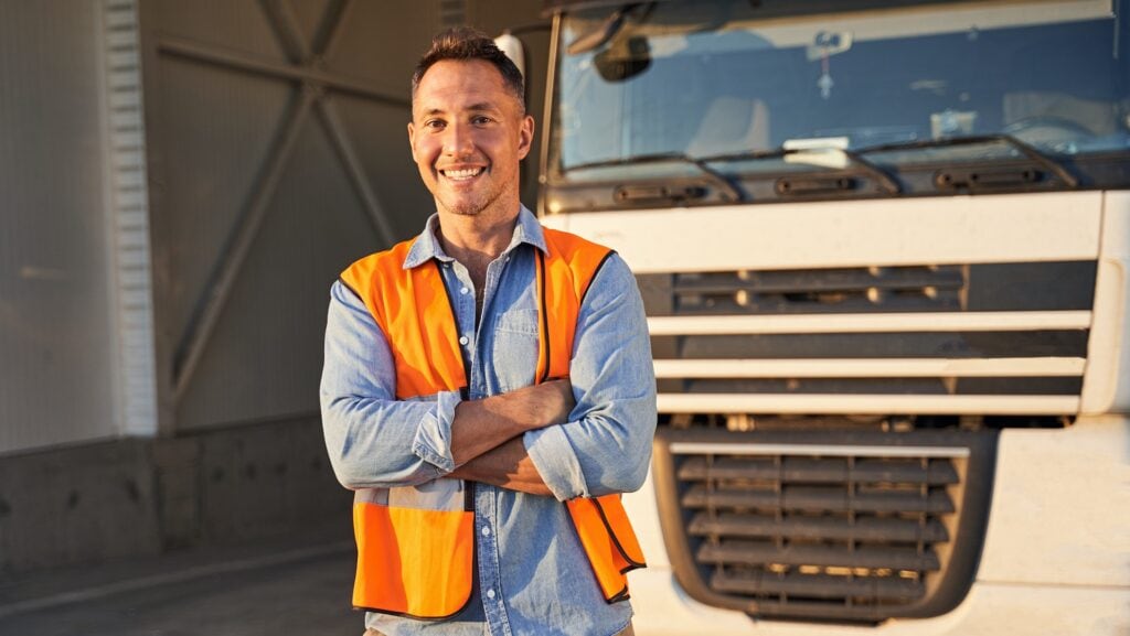 A smiling man in a high-visibility vest standing in front of a large truck, representing driver training at Suffolk New College.