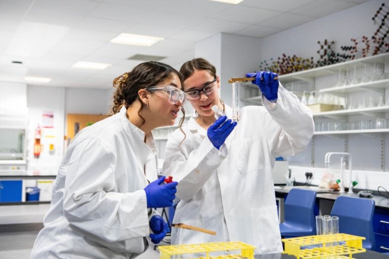 Two students in lab coats and safety goggles conducting a science experiment in a Suffolk New College laboratory, focusing on practical science skills and education.