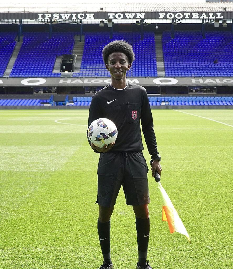 Young man in referee uniform holding a football and flag on the pitch at Ipswich Town Football Stadium, Suffolk, with empty blue seats in the background.