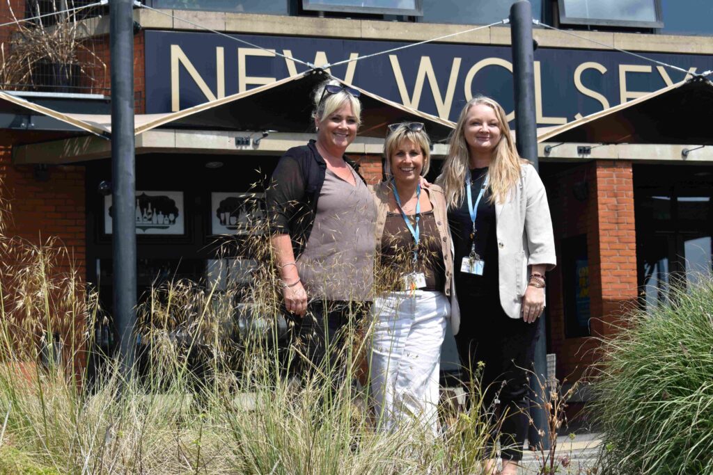 Three women standing outside Suffolk New College theatre, smiling, with a sign reading 'New Wolsey' in the background, representing creative careers.