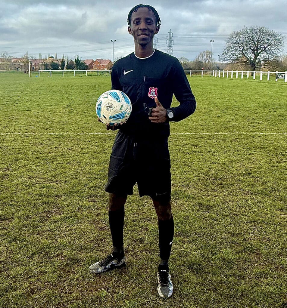 Ipswich man in football kit holding a ball on a football pitch, smiling, with a thumbs-up gesture, under cloudy sky, representing determination and sportsmanship.