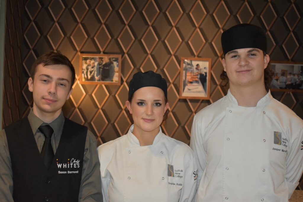 Three Suffolk New College students in chef uniforms standing in a restaurant, smiling, with a patterned wall and framed photos in the background.