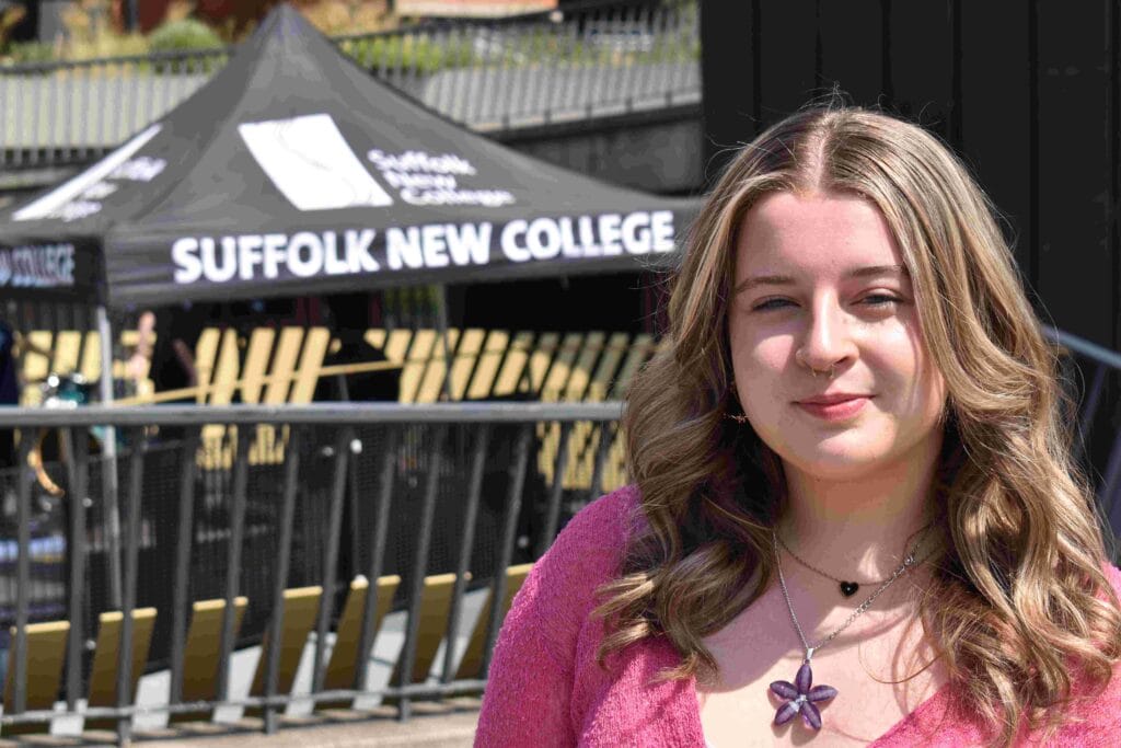 A young female student with long wavy hair, wearing a pink top and butterfly necklace, standing outdoors in front of Suffolk New College theatre tent, showcasing creative careers.