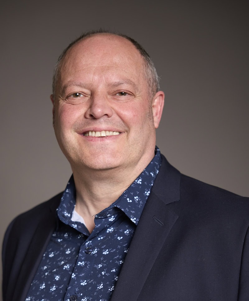 Portrait of Richard Brame, smiling, wearing a dark blazer and patterned shirt, in a professional setting at Suffolk New College for SEO-friendly image description.