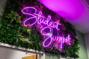 Neon sign reading 'Student Support' illuminated in purple, surrounded by green plants at Suffolk New College, highlighting student assistance services.