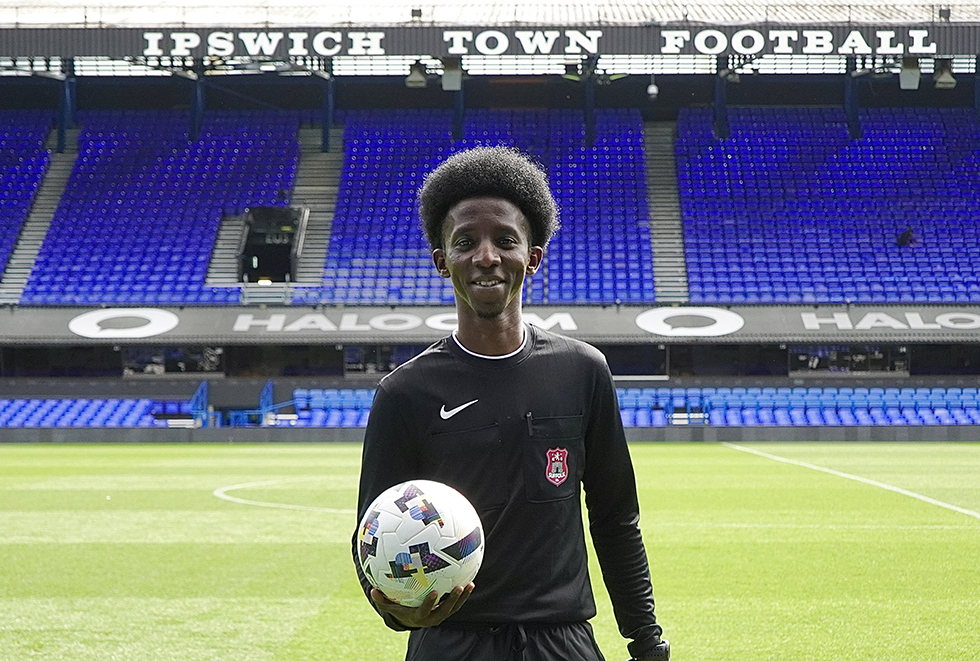 Young Ipswich football player holding a soccer ball on the pitch at Ipswich Town Football Stadium, with empty blue seats in the background.