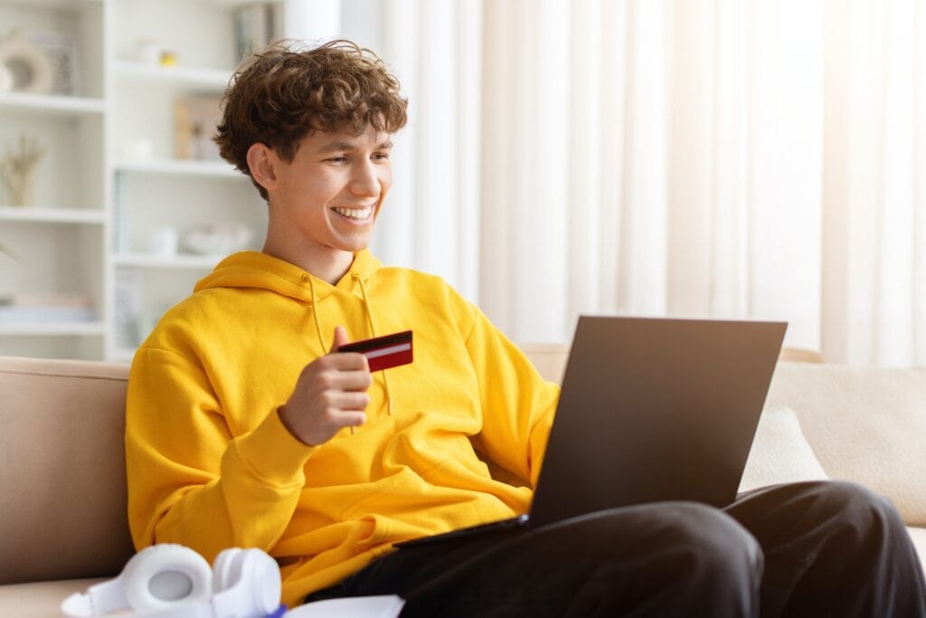 Young male student in a yellow hoodie holding a credit card and using a laptop at home, representing financial support and online learning.