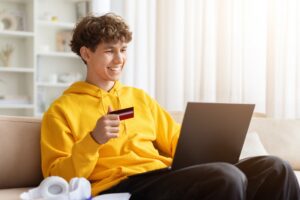 Young male student in a yellow hoodie holding a credit card and using a laptop at home, representing financial support and online learning.
