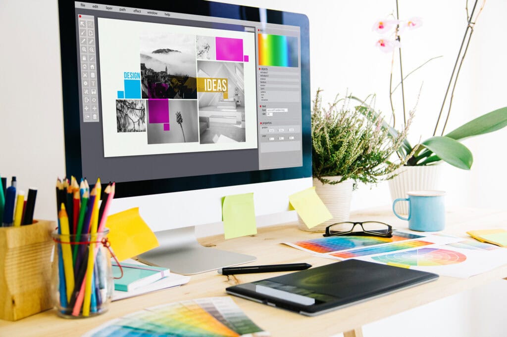 Modern digital design workspace with computer displaying graphic design software, surrounded by colour swatches, stationery, and plants at Suffolk New College.