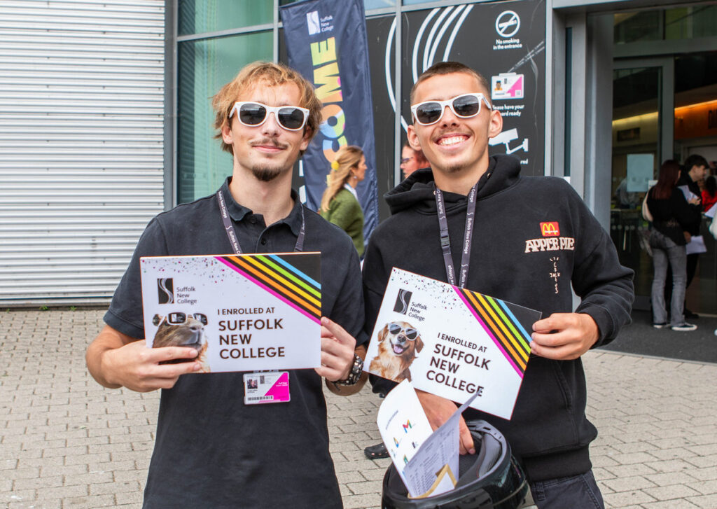 Two smiling students holding Suffolk New College enrolment leaflets outside the college entrance, promoting education and student life.